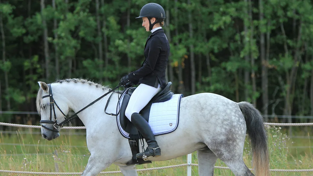 Op de foto: vrouw tijdens een dressuurwedstrijd op een wit paard | De beste paardrijlaarzen met een extra wijde kuit
